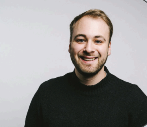 Portrait von Lukas Burnar. Ein junger Mann trägt einen Rollkragenpullover und lächelt in die Kamera.